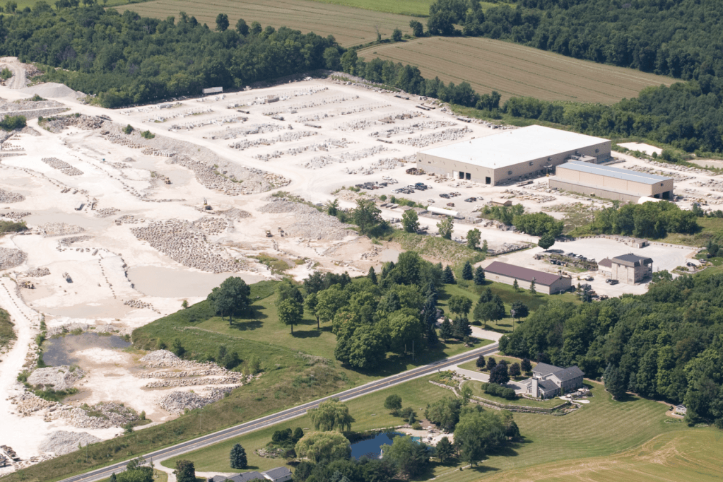 Aerial view of a large industrial site with a processing facility, surrounding rock piles, and a variety of vehicles, set amidst greenery and farmland.