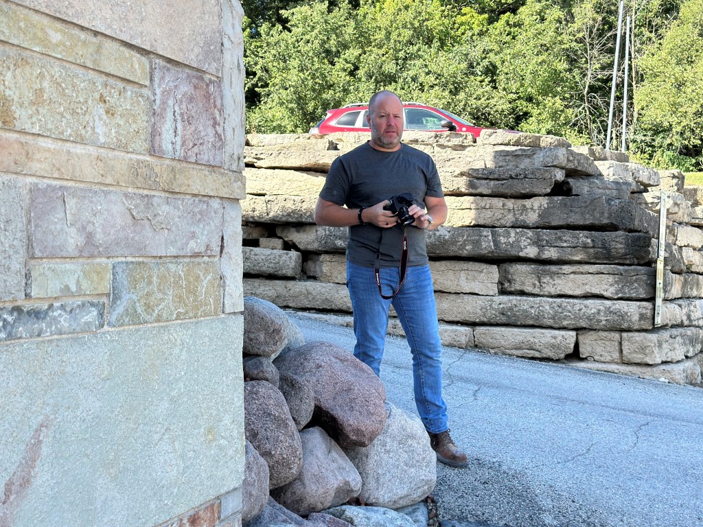 A man holding a camera stands near a stone wall, with a red car parked in the background and trees visible behind.