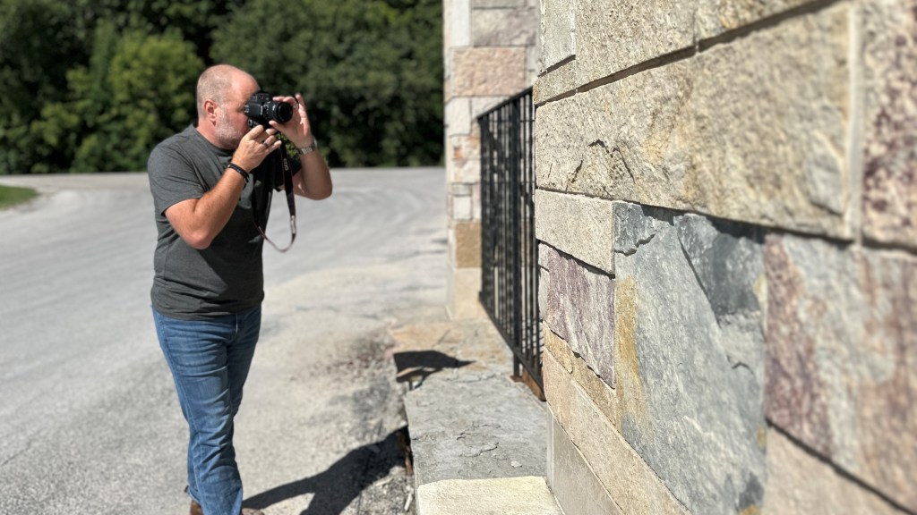A man holding a camera and photographing a stone wall outdoors on a sunny day.
